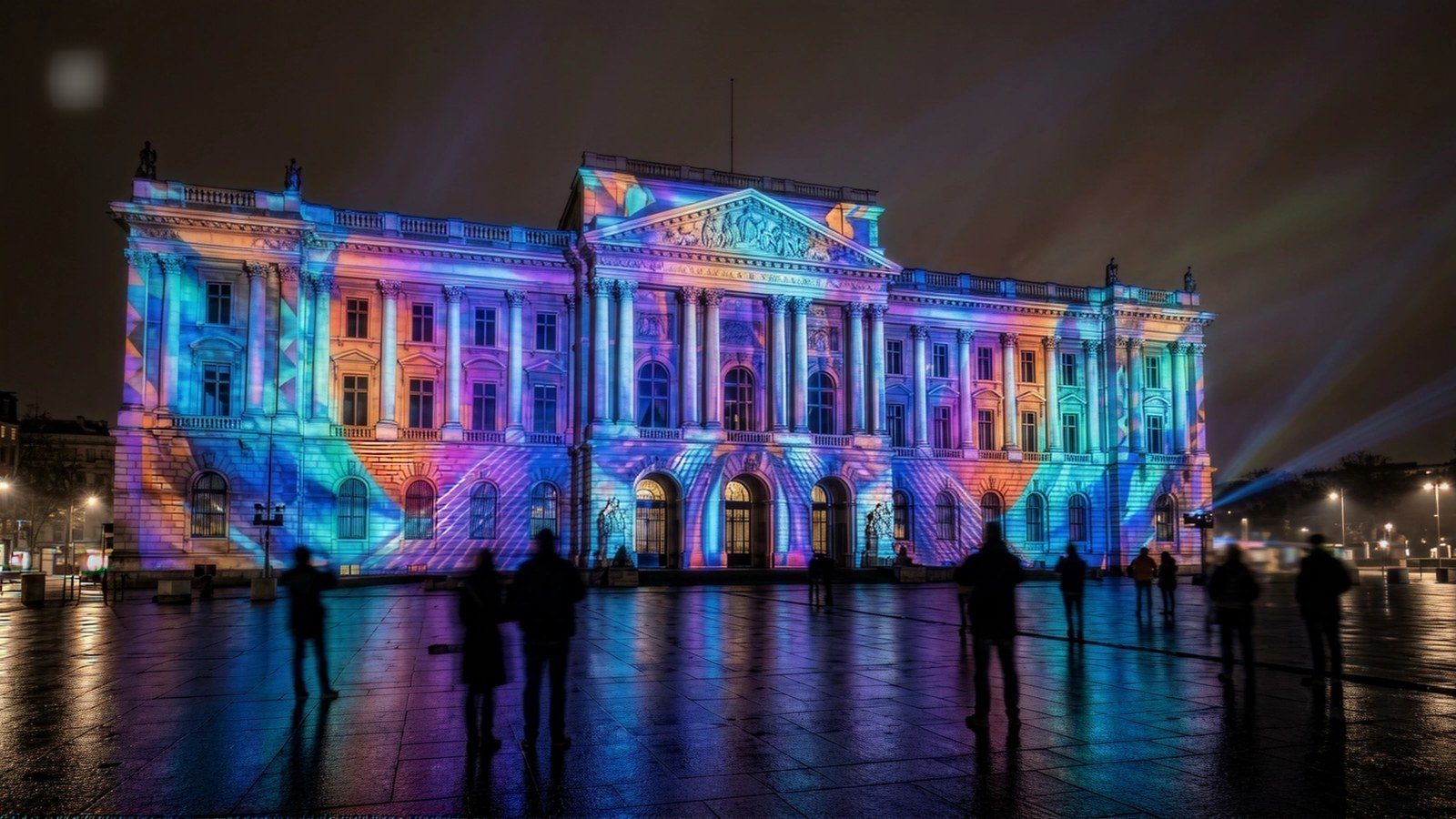 Crowd watching a large projection show on a historic building, reflections in wet ground
